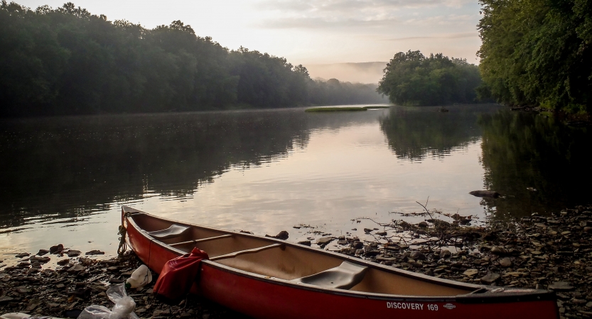 A canoe rests on a rocky shore near a calm body of water framed by trees. 